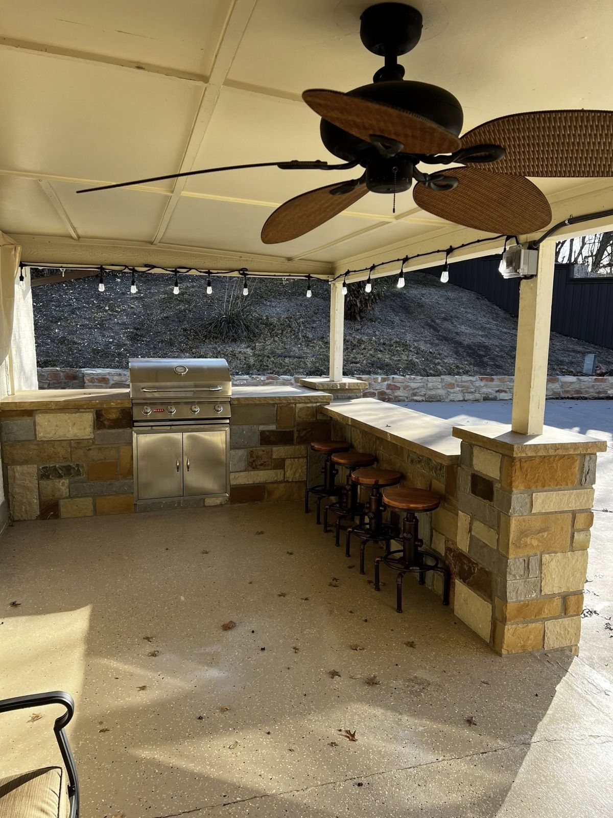 Covered outdoor bar and kitchen with stone counter, stools, and string lights